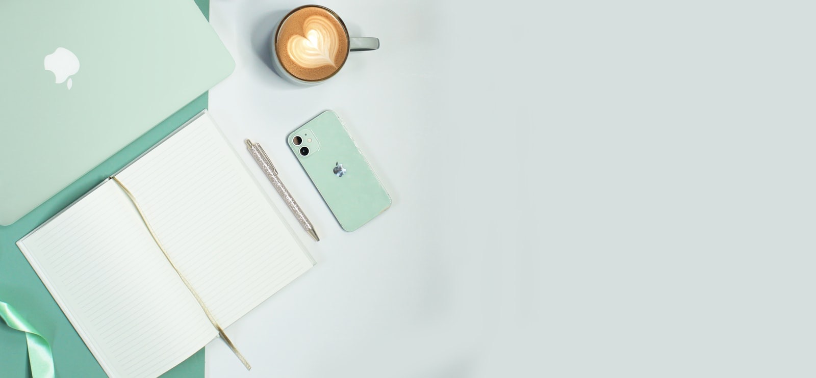 An open notebook, a phone and a cup of coffee laid out on a pale green desk